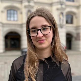 Luna Spiegel mira directamente a la cámara, con gafas de montura oscura y una blusa negra; detrás, la fachada clara y ornamentada de un edificio.